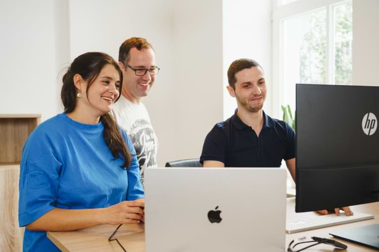 Eine Frau und zwei Männer stehen um einen Schreibtisch mit Laptop und Monitor